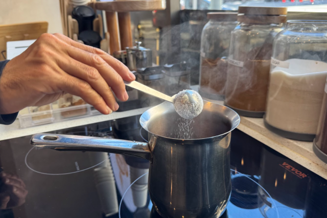 Close-up of a hand holding a measuring spoon, sprinkling a powder into a steaming saucepan on a stovetop, with jars of ingredients in the background.
