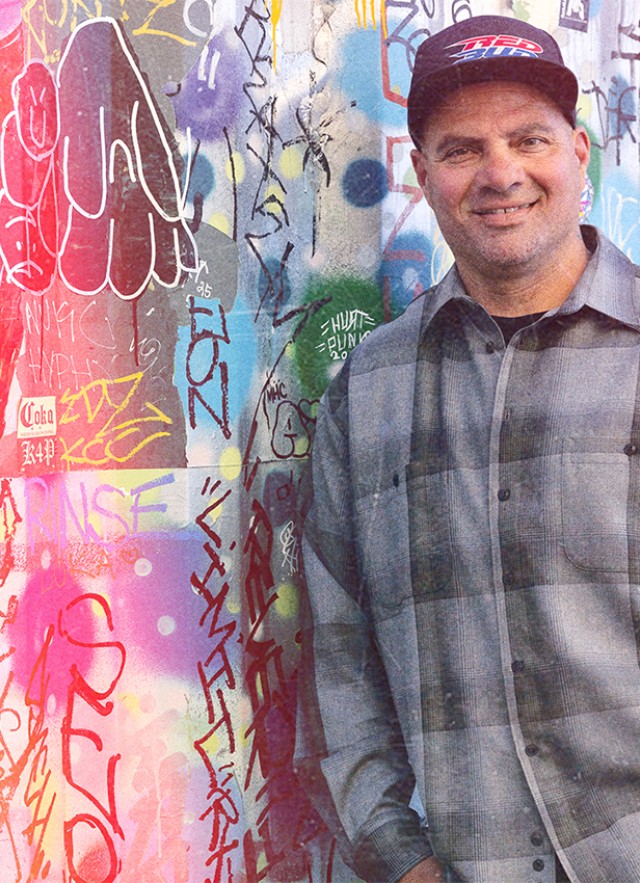 Man standing in front of a call covered in colorful graffiti, leaning his right shoulder against the wall. His hands are in his pockets; he wears a dark baseball cap and gray plaid, button-down long-sleeved shirt.