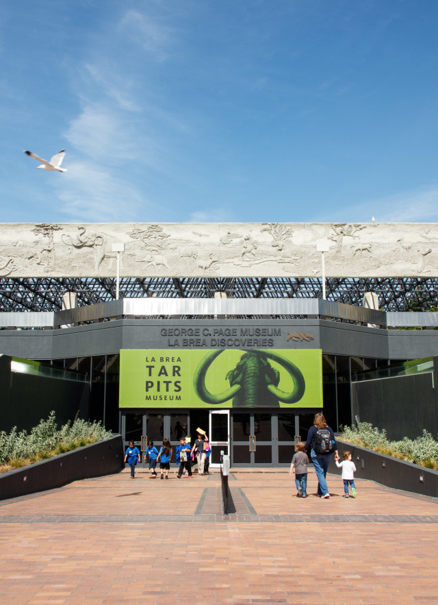 A few people walk towards the entrance of La Brea Tar Pits museum a flat-topped recessed entrance with a green banner featuring a mammoth on it.