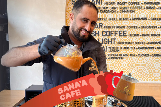 A man in a black vest and gloves smiles while pouring spiced tea from a glass teapot at Sana’a Cafe. The background features a patterned menu of Yemeni coffee and tea, with a red promotional banner in the foreground.