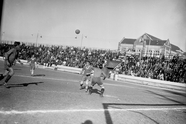 Black and white photo from the 1930s of men playing soccer outside with a packed crowd in bleachers behind them.