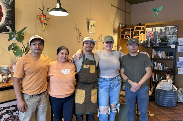 A family portrait of cafe owners Rosa and her son Jefri, standing with Jefri's partner Michaela, uncle Elvis, and brother Christopher. They are smiling in the cafe's cozy seating area, which features warm ambient lighting, vibrant house plants, and artwork on the walls.