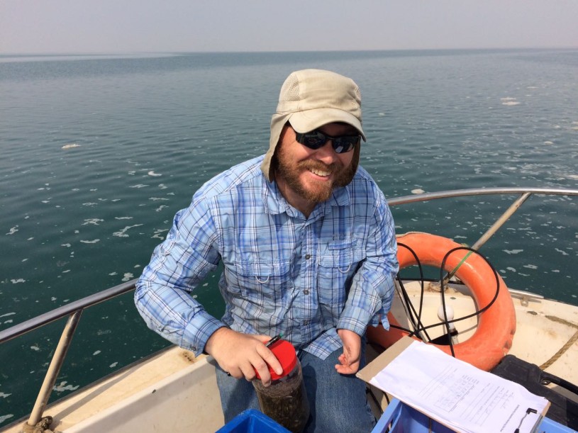 A bearded man in hat, sunglasses, and blue shirt holds a specimen jar and clipboard while sitting in the back of a boat next to an orange lifesaver