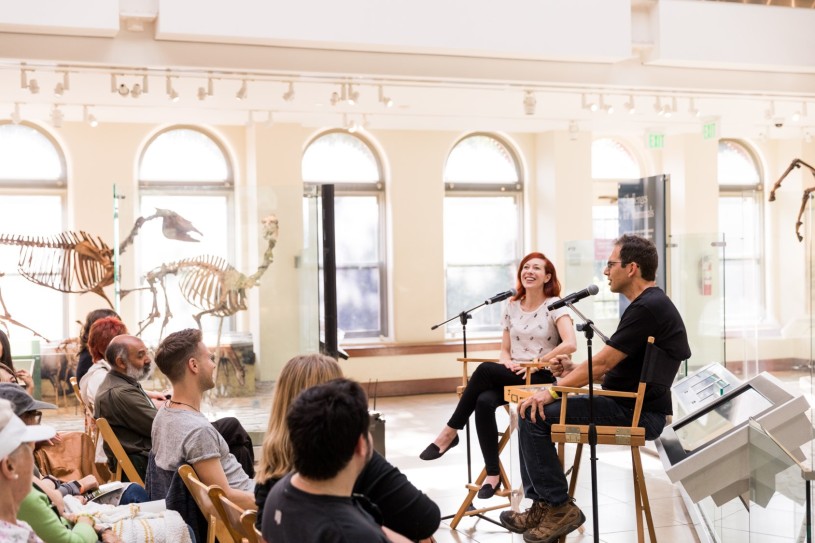 Two panelists sitting in directors chairs, speaking into microphones, in front of an audience listening to their discussion.