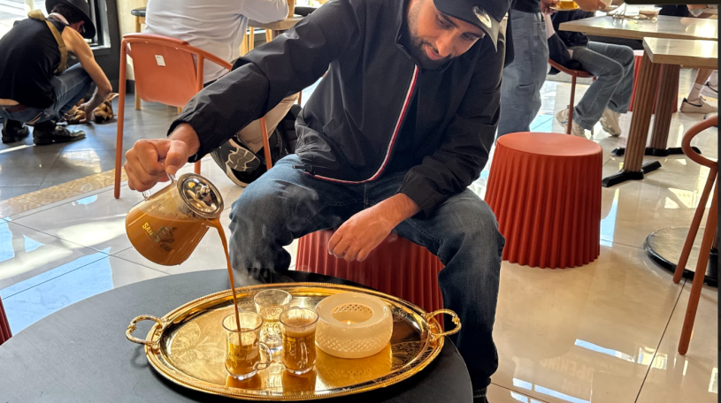 Man carefully pours a warm beverage from a clear glass teapot into a small glass cup, beautifully arranged on a large, ornate gold serving platter.